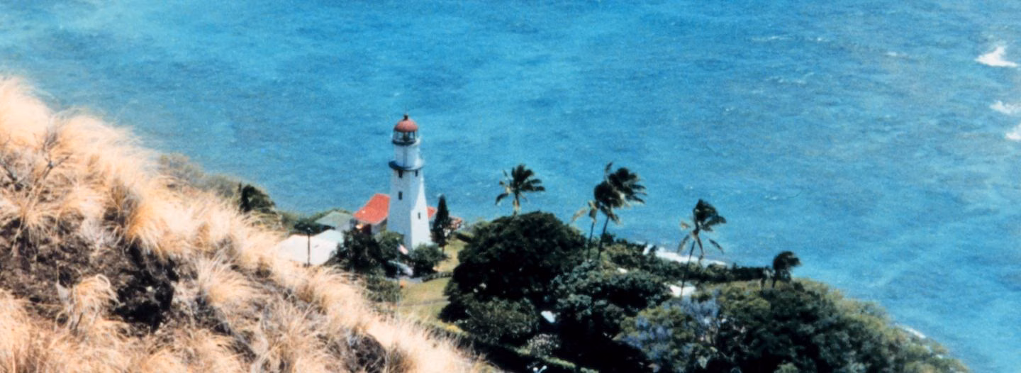 The white Diamond Head Lighthouse with its red roof on the shore below Diamond Head, seen from above the cliff