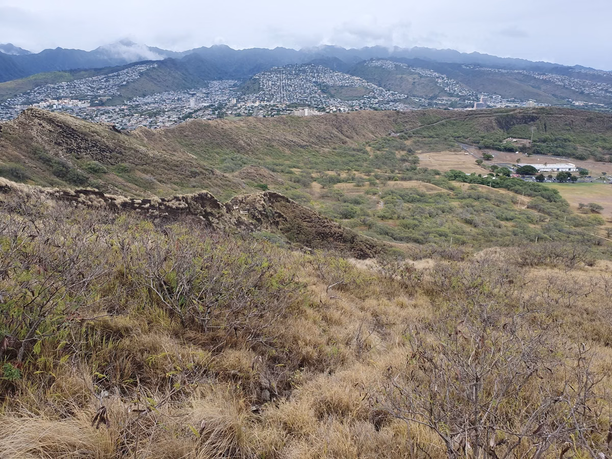 The grassy inside of Diamond Head crater and its rim, seen from the summit trail, with Honolulu neighborhoods and mountains behind